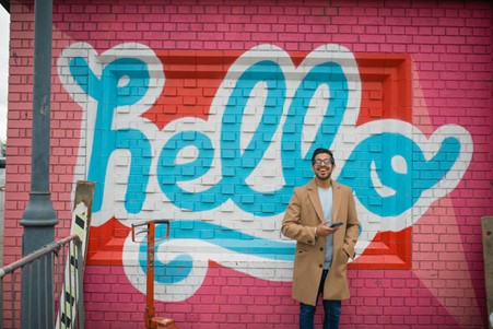 Man stood in front of a pink, white and blue street art sign reading 'hello'