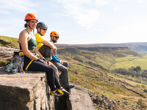 Three people wearing helmets and climbing gear sitting on the edge of a rock up a hill surrounded by greenery