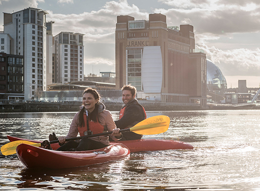 A man and woman smiling in kayaks on a sunny day in a Newcastle river