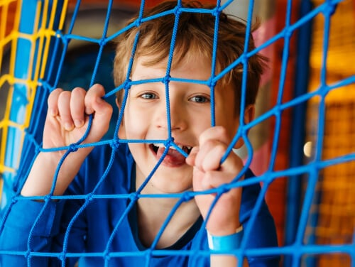 a smiling boy behind a lattice of rope which makes up the walls of a jungle gym. The boy is gripping the lattice, pulling it down slightly.