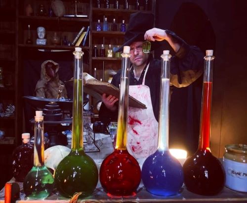 A man in a bloody apron and a top hat looking into some test tubes filled with different liquids.