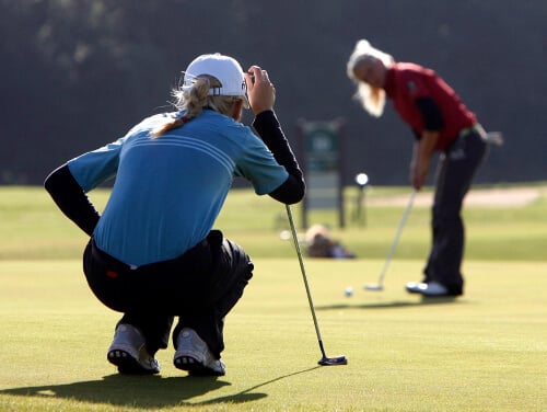 Two women wearing outfits for golf out on a sunny golf course with one crouched down to watch the ball
