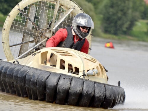 a man in a hovercraft going at considerable speed. He is wearing a helmet and life vest and is currently above water.