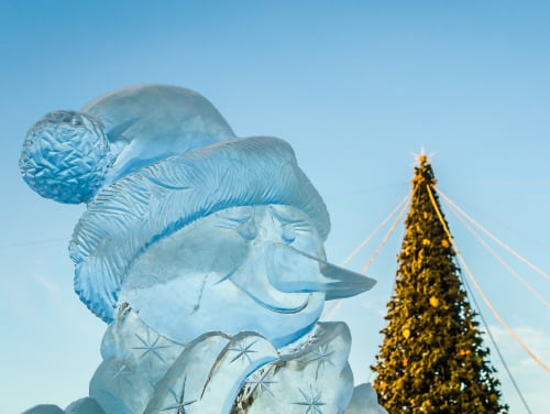 A snowman made of ice with a giant christmas tree behind him fully decorated,