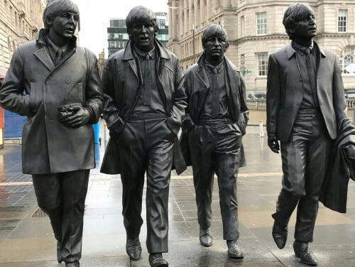4 individual statues of the Beatles looking like their in motion walking somewhere. They all wear smart jackets like pea coats and suit jackets.