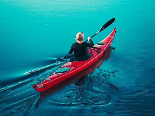 The back of a blonde woman in a red kayak going down a blue river