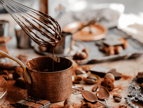 A brass pot filled with melted chocolate with a whisk dripping chocolate into it. 