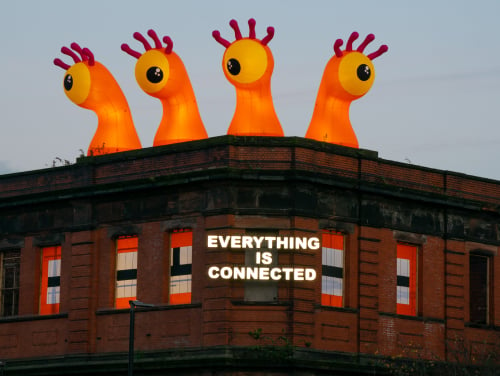 4 eyes of a monster peeking up behind a building in manchester with the words "everything is connected" on the building