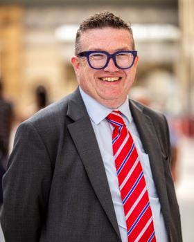 Professional image of Martin Keating wearing a dark suit and red, white and blue stripped tie with glasses