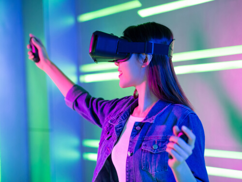 A woman smiling whilst wearing a VR headset, in her hands she holds separate controllers.