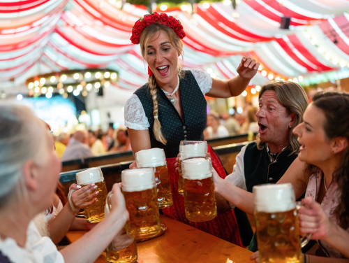 A women dressed in traditional Bavarian clothes handing out steins of beer to a table of smiling adults.