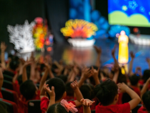 Children with their arms in the air as they are sat in a theatre in front of stage.