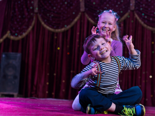 Two kids on a stage playing, a boy sits in front with a girl behind him.