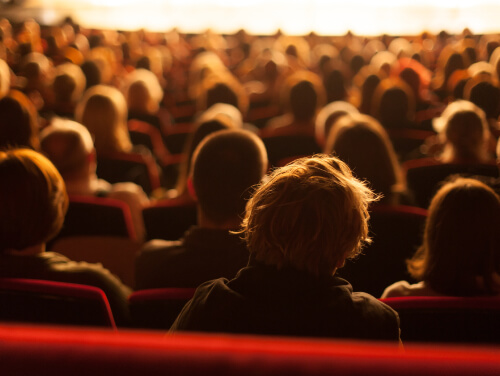 the backs of a crowd of people's heads as they look towards a stage. they are sat in a theatre.