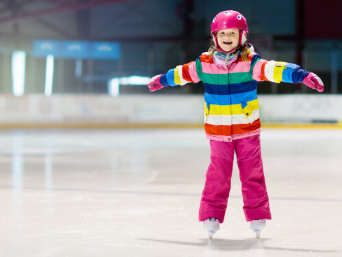 A small girl wearing colourful clothing with her arms outstretched skating on an ice rink