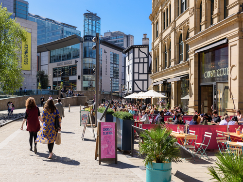 A busy street in Manchester in the sunshine with people sitting outside restaurants