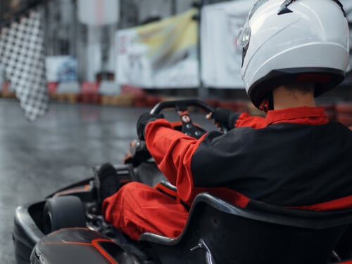 Person wearing a red and black racing suit driving a Go Kart on an indoor racing track.