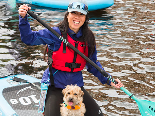 A woman on a paddleboard smiling with a paddle in her hands. There is a dog with her on the board