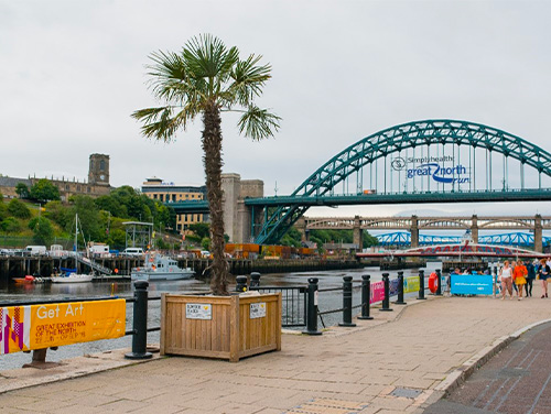 A bridge over the Tyne, there are a couple of boats on the river and people on the banks of the river.