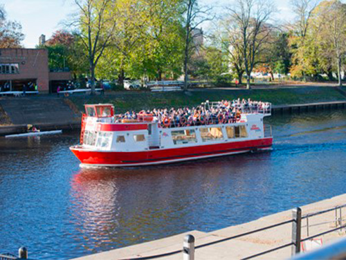 A cruise boat on the River in Ouse in York. The boat is packed with people on its upper deck as it glides through the water.