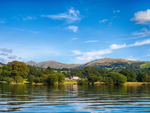 Lake windermere on a sunny day with a house pictured across the water on a bank.