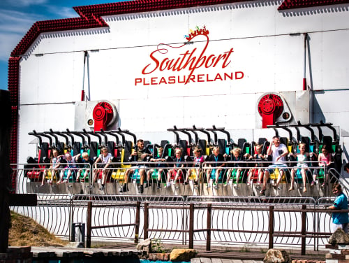 A ride at southport pleasureland with a mix of children and adults on it. The ride will spin them round vertically. It is a sunny day.